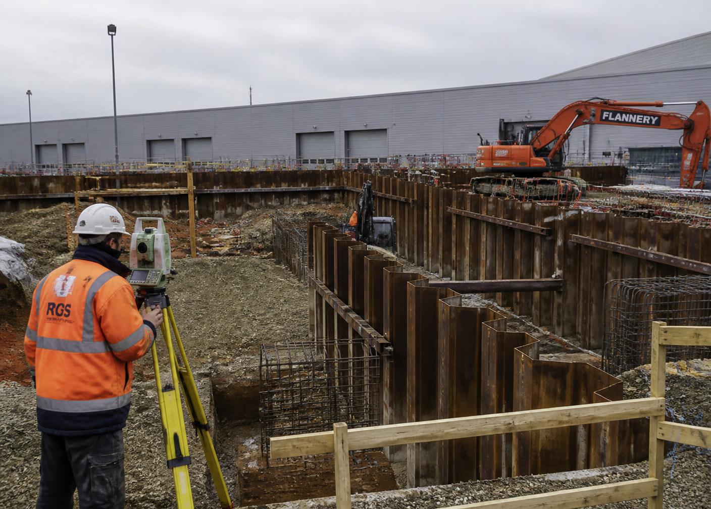 Ironbound Geotechnical surveyor recording elevations beside sheet-pile shoring at a warehouse excavation